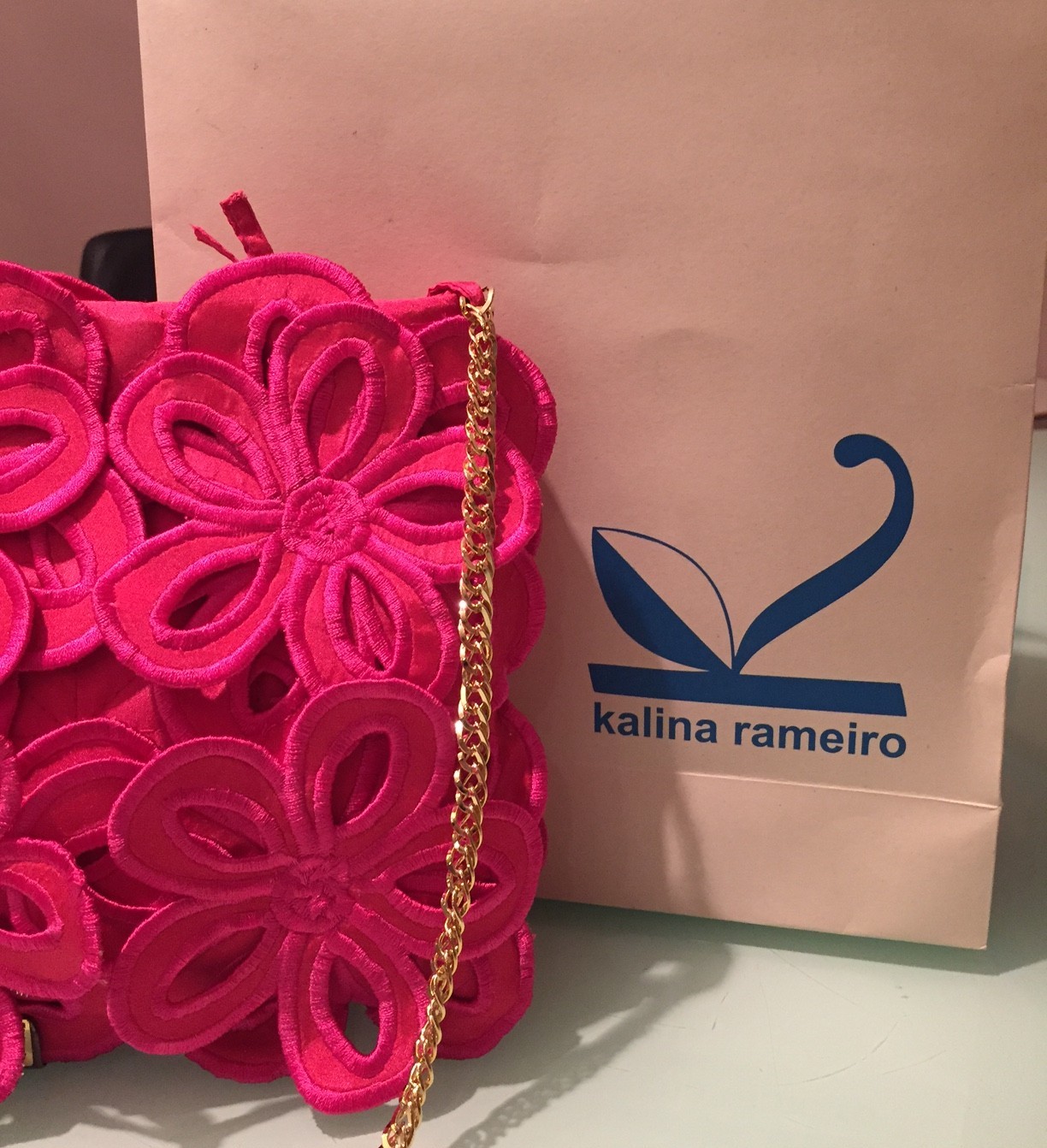 bolsa na cor lilás, em tecido com flores da mesma cor, sobrepostas, a alça é em corrente dourada. logo atrás uma sacola de papel , com os dizeres: Kalina Rameiro.