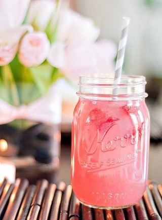 Em um pote de geléia está um suco cor de melancia : vermelho claro com um canudo listrado de cinza e branco. Ao fundo está um vaso de rosas cor de rosa desfocado.