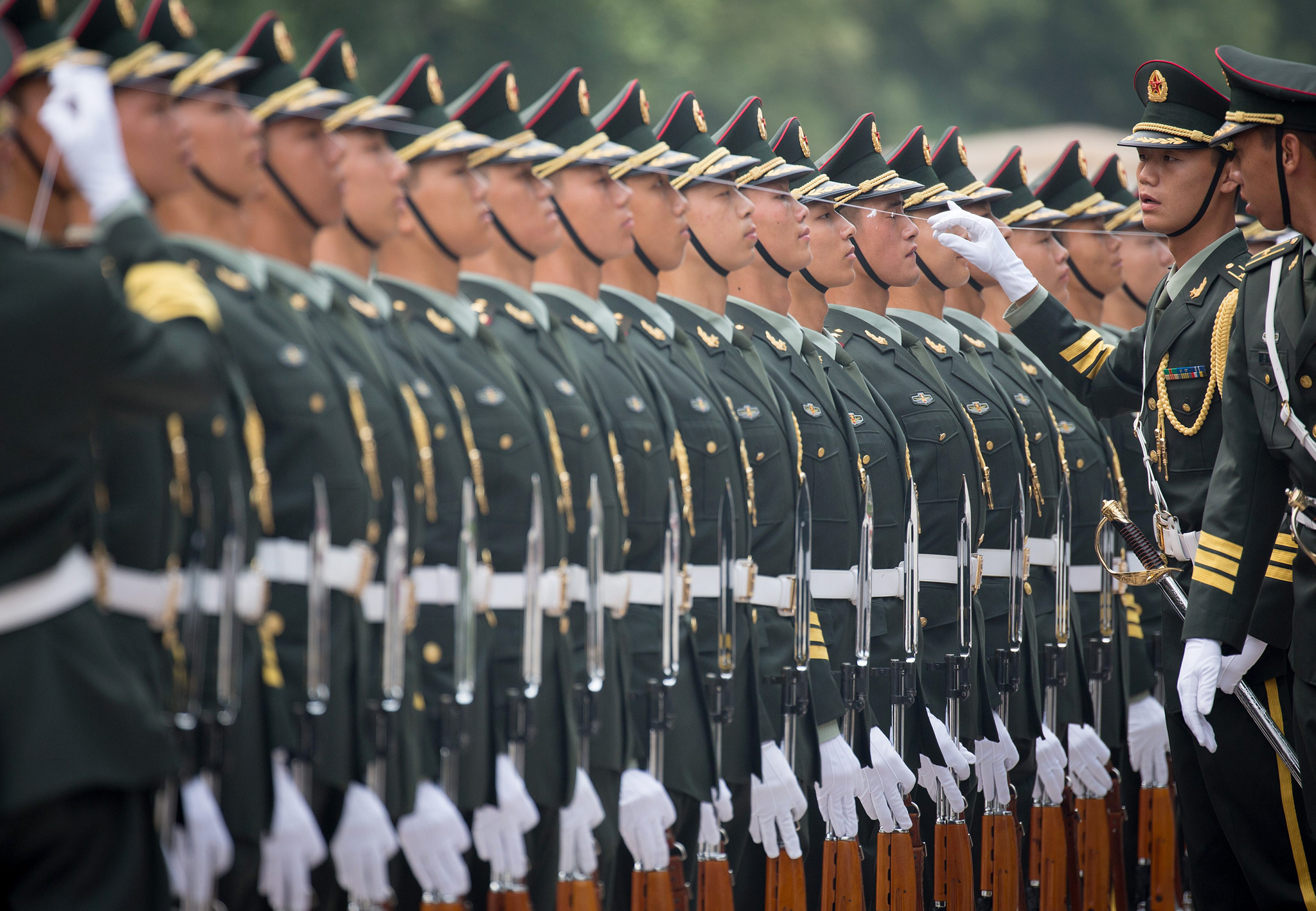 Tropa Militar Chinesa em formação. todos de uniforme verde escuro. usam quepe da mesma cor. Observa-se a perfeição do alinhamento , pois eles usam uma linha, barbante que nivela o quepe na cabeça de todos eles. Um oficial comandante da tropa observa esse alinhamento.