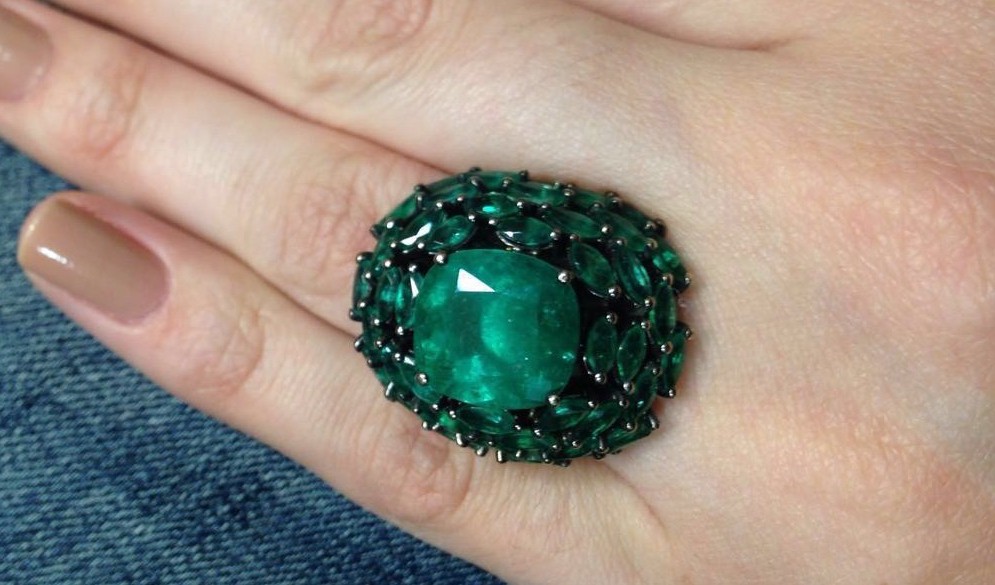 uma mão de mulher está em close usando um anel de pedra esmeralda verde oval rodeado por três fileiras de esmeraldas pequenas em formato de losango. A aparência é de uma jóia antiga e muito valiosa