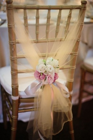 Uma cadeira em madeira está enfeitada com um delicado véu em tom creme que está arrematado em seu encosto com um pequeno buquê de rosas brancas e cor de rosa amarrando todo o arranjo com fitas de cetim no mesmo tom.