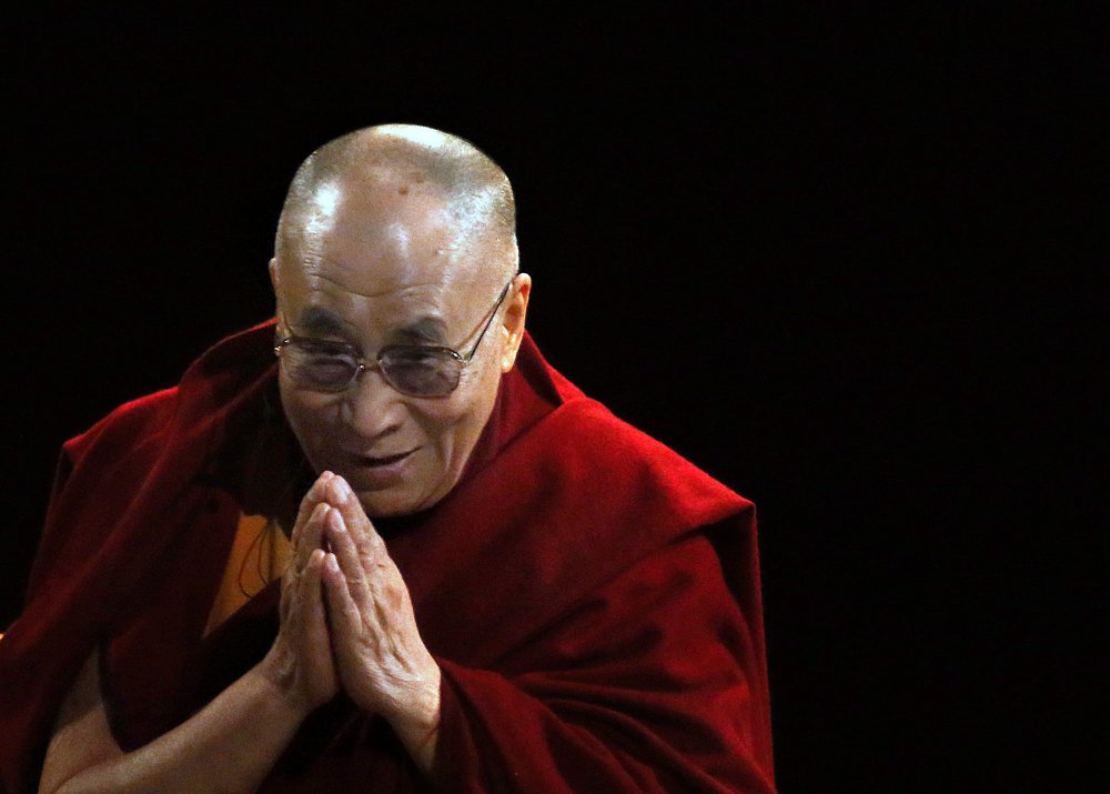 Venerável Dalai Lama, líder religioso do Tibet, num ambiente escuro, usa as túnicas marsala, símbolos dos monges tibetanos, as mãos unidas em mantra de oração. Ele tem o sorriso da compaixão. Ele é careca e usa óculos.