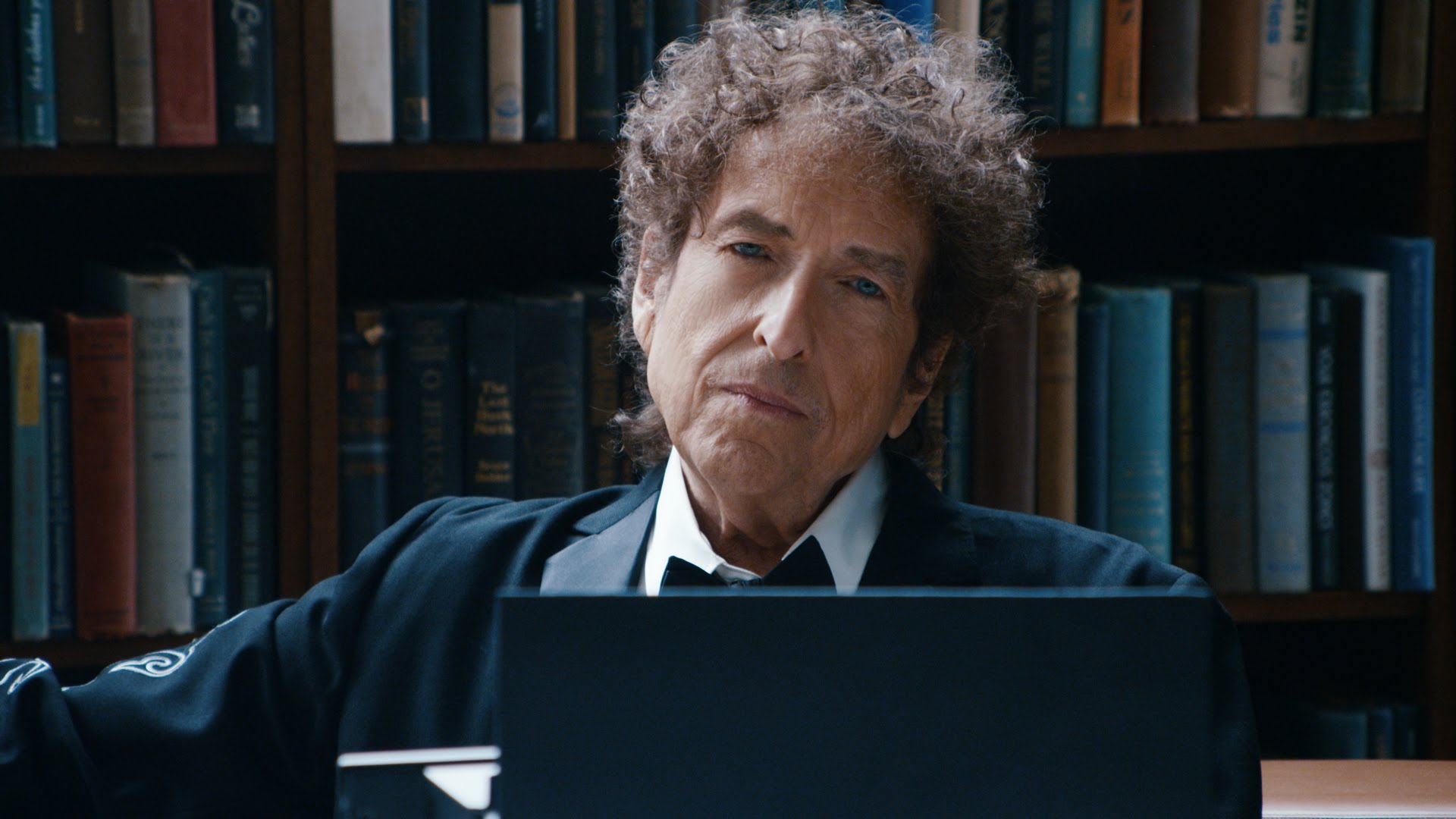 Cantor americano Bob Dylan em foto numa biblioteca , onde aparece seu rosto , cabelos desarrumados, rosto esguio, camisa branca e casaco preto. nariz afilado, olhos claros. Seu semblante é de contemplação de algo.