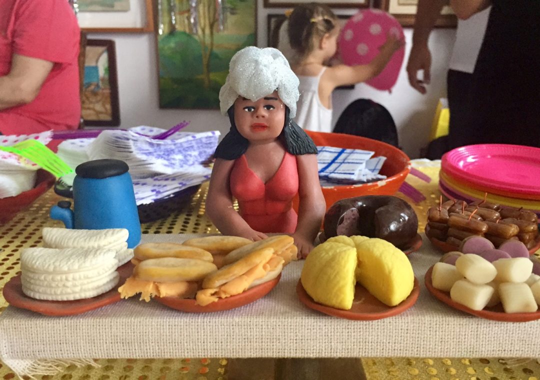 Mesa de festa, com pequenos pratos, com várias comidas típicas brasileiras. e ao centro tem uma imagem das namoradeiras , em barros.