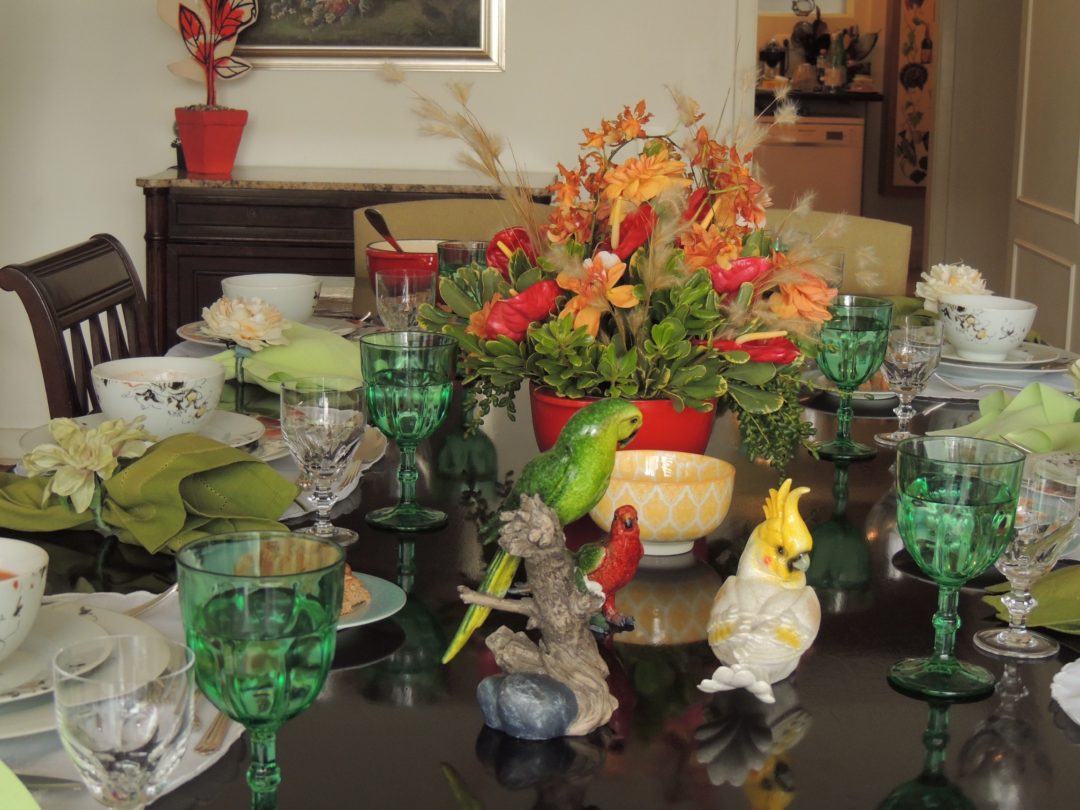 Mesa de jantar montada , com todos os utensilios e acessórios. Flores ao centro na cor branca e laranja. Algumas taças estão em detalhe - a taça de água em tom verde e a taça de vinho branco, em cristal transparente. Como decoração muitos pássaros, araras, papagaios em porcelanas estão em volta do vaso de flores