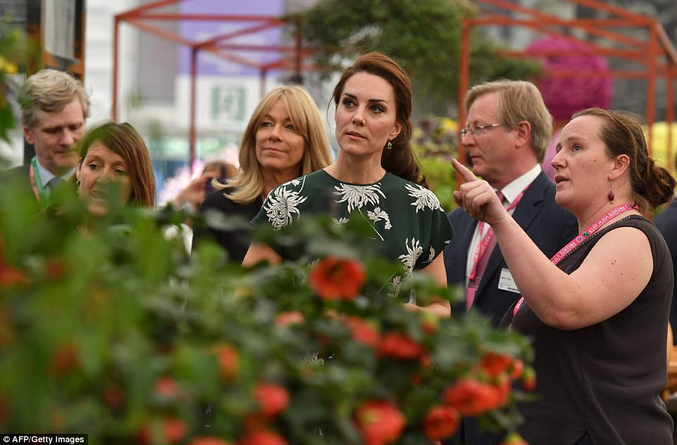 Kate Middleton, em visita a Chelsea Flowers Show, sorri para todos, usando um vestido verde escuro e com detalhes de flores na cor branca, tem seus cabelos presos, Ela está olhando a exposição junto a um grupo de pessoas.