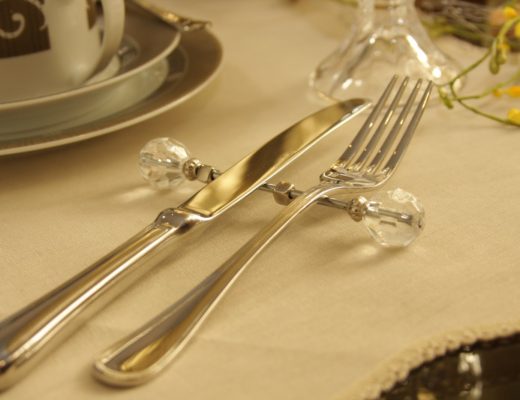 talheres em prata, garfo e faca, apoiados num descanso fino, em prata, com bordas em vidro transparente.