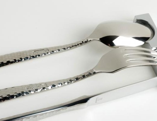 Uma colher, um garfo e uma faca de prata, estão apoiados num descanso de talheres em forma de encaixe geométrico de cada um deles.