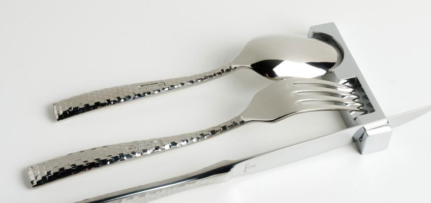 Uma colher, um garfo e uma faca de prata, estão apoiados num descanso de talheres em forma de encaixe geométrico de cada um deles.