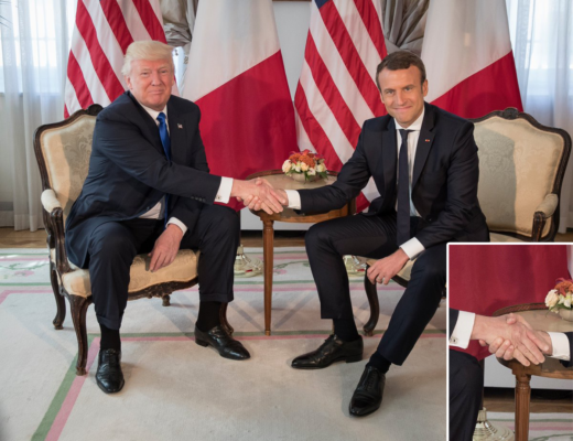 Presidente Donald Trump e o Presidente Emmanuel Macron da França, participam do Encontro Protocolar, eles estão sentados um de cada lado, com bandeiras dos dois países ao fundo, e Macron aperta a mão de Trump, fortemente, ambos vestem ternos escuros , camisas branca e gravatas em cores escuras