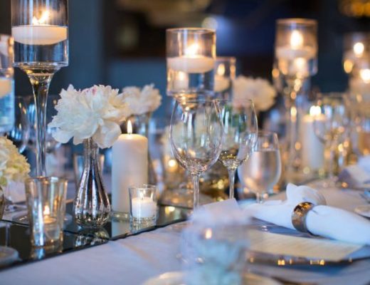 Mesa de jantar com muitos objetos de decoração, tudo em tons claros, alguns em branco. muitas velas na base ao centro e outras velas em castiçal em vidro com água .