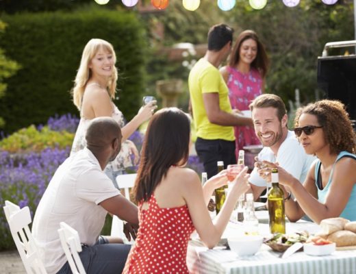 Num jardim, pessoas participam de um almoço. Á mesa dois casais brindam com taças de vinho , enquanto outras pessoas estão em pé junto a outra mesa.