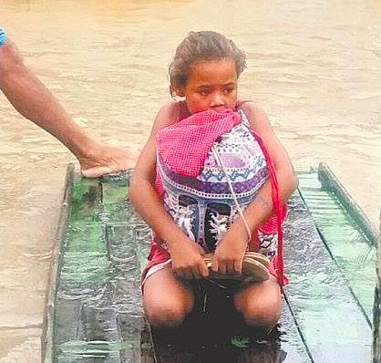 Na enchente em Pernambuco, menina de 8 anos, sobre uma tabua de madeira, que serve como barco, está ajoelhada com uma mochila abraçada ao corpo e dentro dela, seus livros e cadernos.